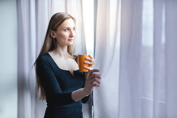 Frau steht mit Kaffee allein am Fenster während der Covid-19 Ausgangssperre