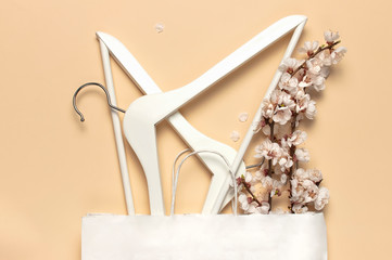 Creative spring sale concept. White wooden hangers with spring flowers white paper bag on beige background top view flat lay. Fashion spring discounts shopping sale store promo design minimalism style