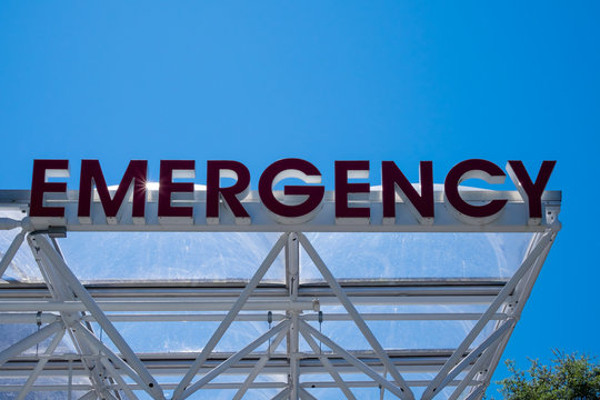A medical hospital's emergency department sign made with red glass. The sign is attached to a clear glass building overhang. There are trees and blue sky in the background. The sign is not lit up.