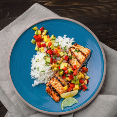 Grilled salmon fillet with fruit sauce made of mango, bell pepper, onion and avocado with basmati rice garnish on a blue plate. Delicious seafood meal. Top view shot above.