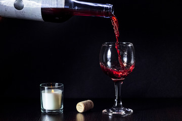 Red wine pouring from the bottle to the glass with splashes and candle on black background