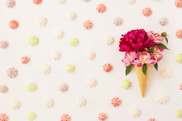 Beautiful fresh peonies in ice cream waffle cone on white background with colored meringue. Stylish flat lay with spring flowers and sweet dessert. Floral card design, copy space.