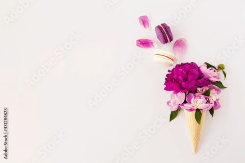 Creative flat lay with ice cream waffle cone with pink and violet peony flowers and macaroons dessert on white surface. Beautiful botanical greeting card or postcard design, empty space for text