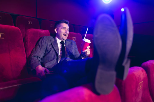 Portrait of a young, handsome man alone in a movie theater in a business suit, with his feet on the front seat and drinking through a tube from a red Cup. - Powered by Adobe