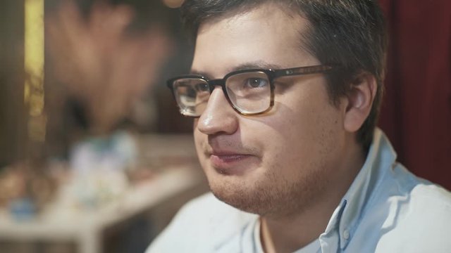 Close up of casual young man in eyeglasses drinking a cup of matcha, in a coffee shop with blurred background. Gimbal shot of man smiling drinking coffee