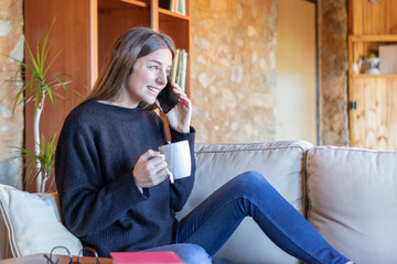 Smiling young brunette woman talking on the mobile sitting on the sofa at home drinking a tea wearing black sweater and blue jeans