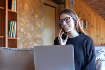 Smiling young brunette woman talking on the smartphone in front of the laptop sitting on the sofa at home wearing black sweater and glasses