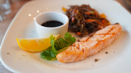 Close-up of delicious dish of buckwheat noodles with salmon and soy sauce with slice of lemon on white plate.