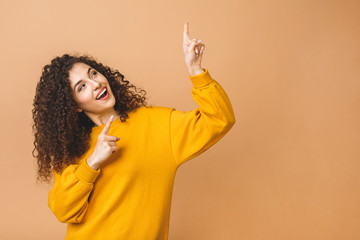 Image of beautiful cute curly young woman standing isolated over beige background pointing up....