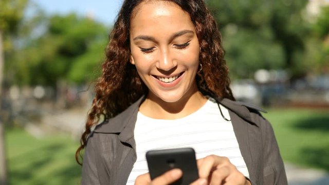 Front view of a happy mixed race woman using smart phone walking in a park a sunny day