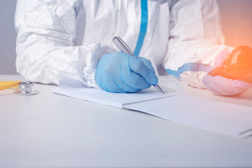 The doctor, who is sitting at the table , in a protective biological suit and gloves , holds an...