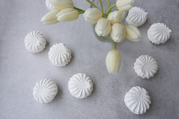 White tulips bouquet in glass vase and sweet marshmallow zephyr, zefir cookies on concrete surface.  Spring bouquet of fresh tulip flowers  and large white meringues on grey table, top view..