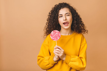 Surprised curly girl eating lollipop. Beauty Model woman holding pink sweet colorful lollipop candy, isolated on beige background.