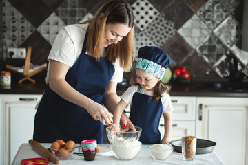 Mother and daughter preparing a sweet cake using flour, milk, sitting on chairs at a table in a modern kitchen. Girl holding a whisk, stirring eggs in a bowl, preparing pancake dough with her mom.