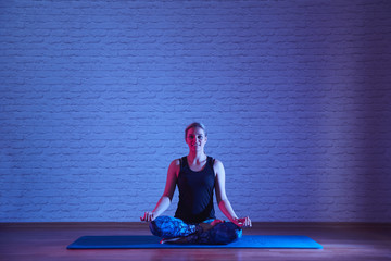 Smiling young woman meditating in lotus position on a yoga mat indoors isolated on wall background