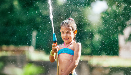 Cute little girl sprinkling with water for herself from the hose, making a rain pleasure for hot summer days. Careless childhood concept image.