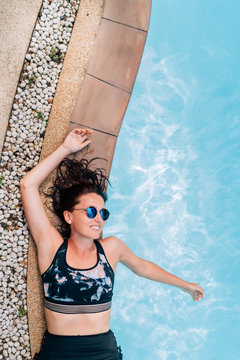 Beautiful sincerely smiling woman in blue sunglasses lying on the Curved swimming pool paved edge with Clean transparent pool water waves background. ..