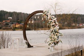 Wooden arch for wedding ceremony with beautiful flowers and retro lamps on the lake, autumn, wedding decor