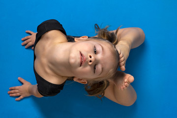 little girl in a black gymnastics leotard does an exercise ring on a blue gymnastic mat, top view