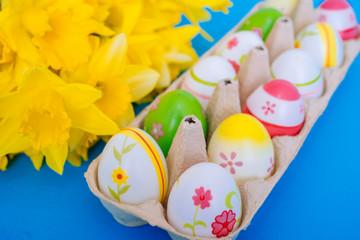 Fototapeta premium Happy Easter. Easter holiday decorations on trendy colored classic blue background. Creative spring composition with Easter elements - flowers and eggs in eggbox. Celebrating Easter concept at spring.