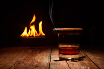 Whiskey and steaming cigar on the wooden weathered table in front of a fireplace.