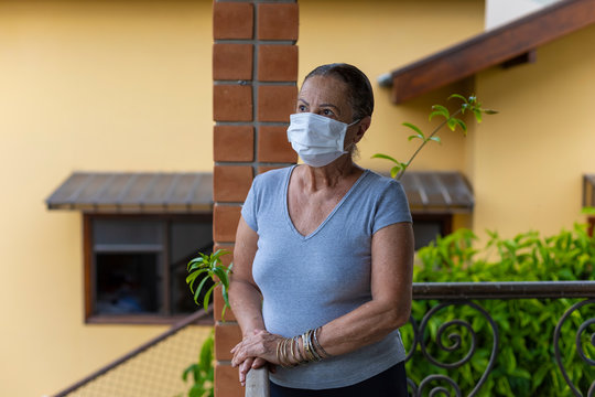Mulher idosa com máscara de proteção e apoiada na varanda de uma casa