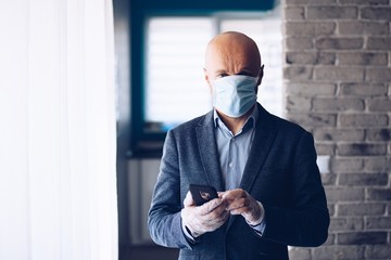 Businessman wearing protective medical mask and using smartphone at home.