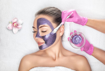 Beautician makes a face mask with purple particles on the face of a woman to rejuvenate the skin. Cosmetology treatment of problem skin on the face and body. - Powered by Adobe