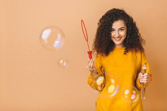 Beautiful brunette curly student girl wears casual making soap bubbles isolated on beige background.