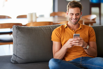Young man texting on his living room sofa and smiling