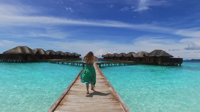 Maldive island resort. Girl in green dress walking on wooden bridge to bungalow. View of a young woman from the back. Maldives travel destination. Summer vacation in paradise. Crystal blue ocean.