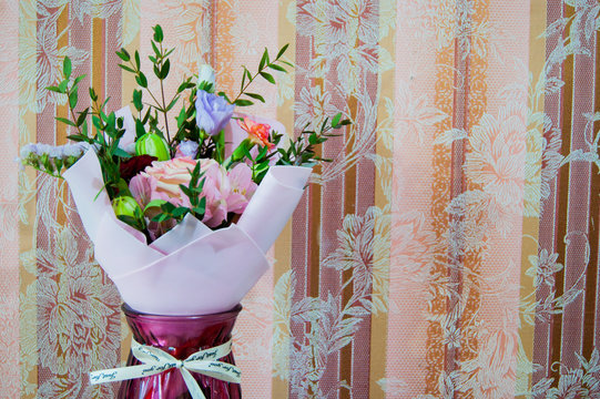 A delicate bouquet of different flowers in a light pink wrapper stands against a background of pink wallpaper in a home setting.