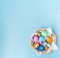 Golden, pink, blue, green, orange eggs in the wicker basket on the blue background. Copy space. Place for text and design. Happy Easter. Top view. Flat layout.