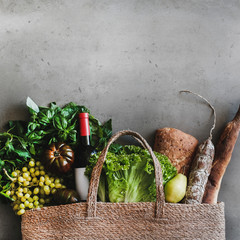 Flat-lay of healthy grocery shopping eco-friendly bag with vegetables, fruit, greens, bread, sausage, wine bottle over concrete background, top view, copy space, square crop. Local farmers market