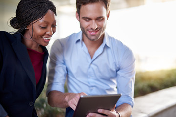 Smiling diverse businesspeople sitting outside working on a tablet together