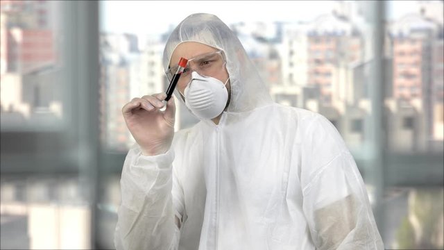 Medical worker in white protective clothing and mask holding test tube with blood. Windows with cityscape on background.