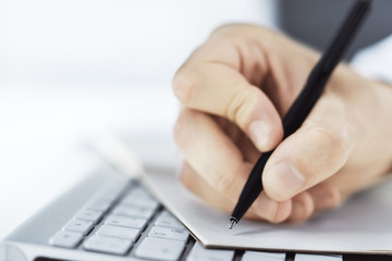 Man writes with a pen in notepad on computer keyboard in a sunny office, business and education concept. Close up