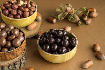 Milky and dark chocolate covered hazelnuts in bamboo bowls on wooden table with natural hazelnuts.