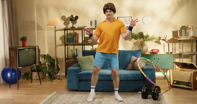 Handsome funny Caucasian retro style man with mustache dancing at blue sofa in room with hula hoop at home. Joyful smiling athlete male model fooling around while posing to camera indoors. 50s concept