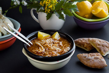 Traditional Turkish Vermicelli Chicken Soup, in the black color bowl with slices of lemon on black surface with chicken legs,bread,lemon and fresh spring flowers.