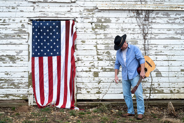 Man wearing cowboy hat and hold acoustic guitar standing in front of old barn with American flag