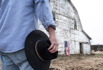 Fototapeta na wymiar Closeup of man holding black cowboy hat standing in front of aandoned old barn on farm with American flag