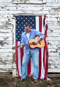 Man Playing Acoustic Guitar Standing On Front Of American Flag Haning On Old Barn Door