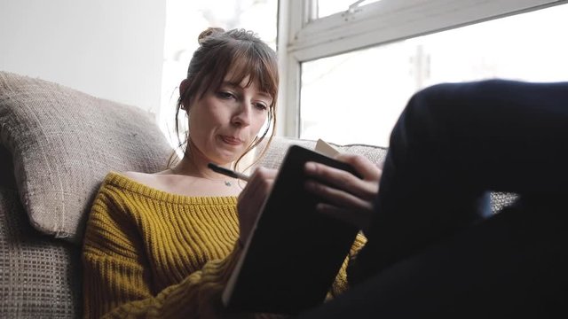 Slow motion shot of woman sitting on couch and writing into a book