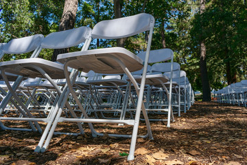 Empty white plastic chairs set up an outdoor in a row for ceremony, event or party under mature trees canopy shade