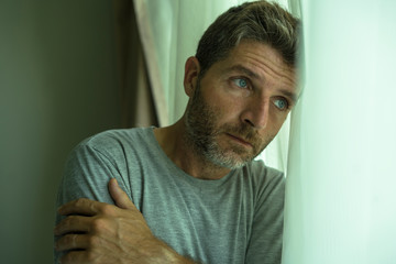mid adult man crying sad and depressed standing at bedroom window in despair at home feeling...
