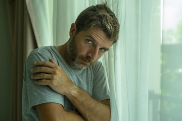 dramatic indoors portrait of mid adult man crying sad and depressed on bedroom window curtain in...