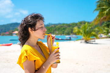 Beautiful girl in a yellow dress drinks fresh mango on the beach of a paradise island. Perfect vacation.
