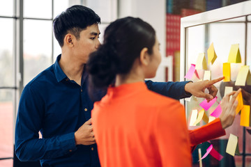 Casual creative happy two smile asian business people planning strategy analysis and brainstorm putting post it stickers note to share idea for startup project on glass board at modern office