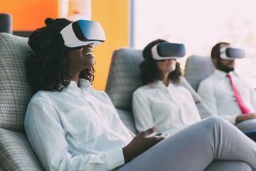 Amazed businesspeople watching virtual presentation. Young black business woman and her colleagues wearing virtual reality headsets, sitting in motion chairs and smiling. VR studio concept